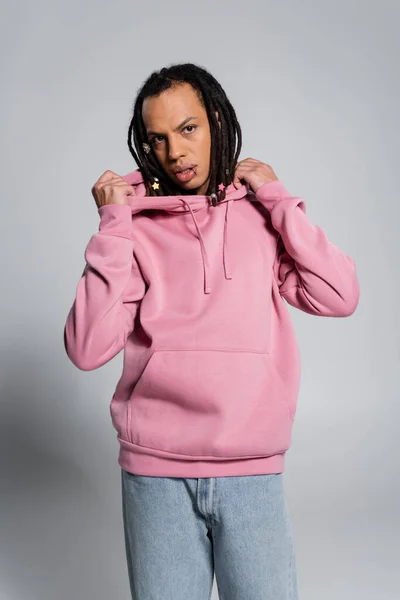 Pierced multiracial man with dreadlocks adjusting pink hoodie on grey - foto de stock