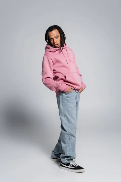 Full length of multiracial and pierced man with dreadlocks posing with hands in pockets on grey — Stockfoto