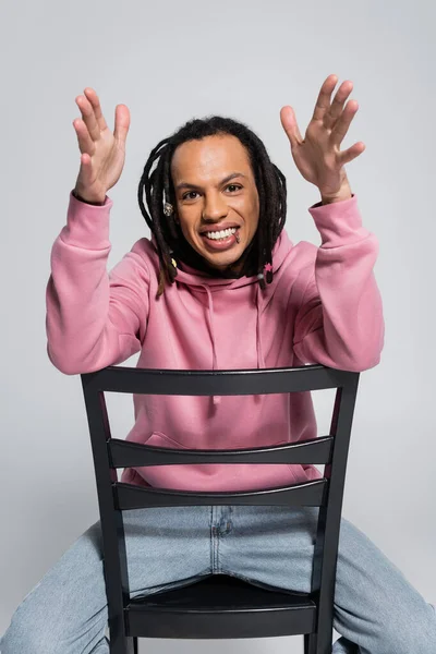 Happy multiracial man in pink hoodie gesturing while sitting on chair isolated on grey — Foto stock