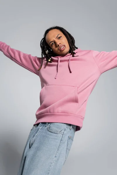 Multiracial man in pink hoodie and jeans looking at camera while standing with outstretched hands on grey — Stock Photo