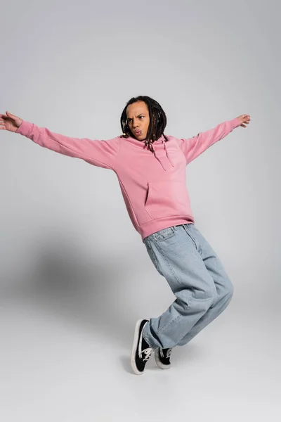 Full length of multiracial man in pink hoodie and jeans posing with outstretched hands on grey — Stock Photo