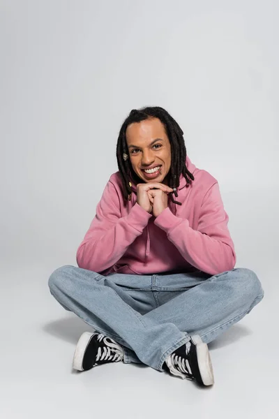 Full length of happy multiracial man with dreadlocks sitting with crossed legs on grey — Stock Photo