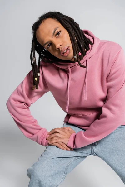 Multiracial man in jeans and pink hoodie looking at camera while posing on grey — Stock Photo
