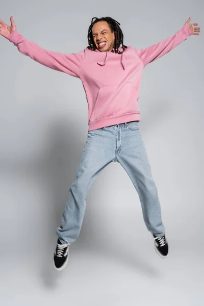 Full length of excited multiracial man with dreadlocks jumping with outstretched hands on grey — Stock Photo