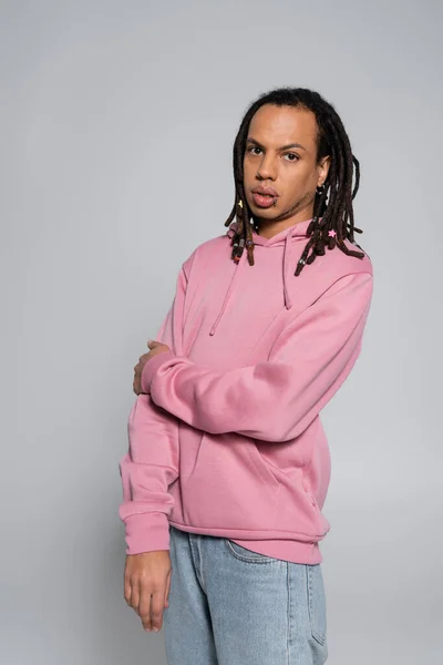 Multiracial man with dreadlocks and piercing standing in jeans and pink hoodie isolated on grey - foto de stock