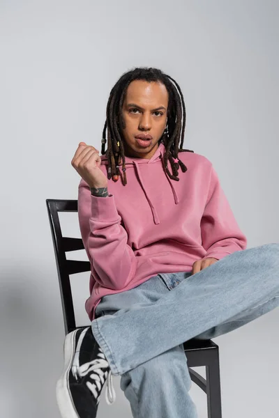 Tattooed and pierced multiracial man with dreadlocks sitting on wooden chair on grey — Stock Photo