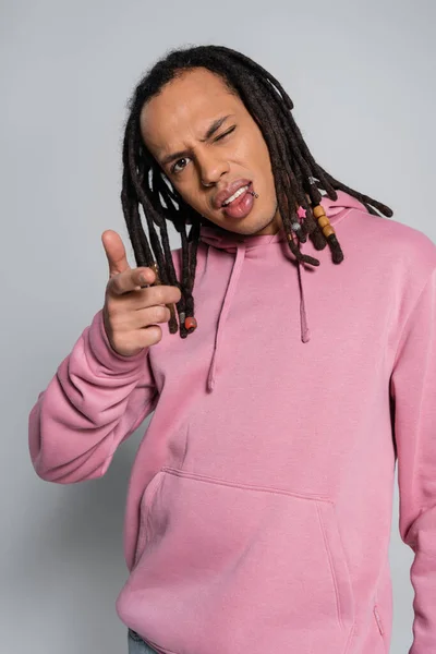 Pierced multiracial man with dreadlocks looking at camera and pointing with finger on grey - foto de stock