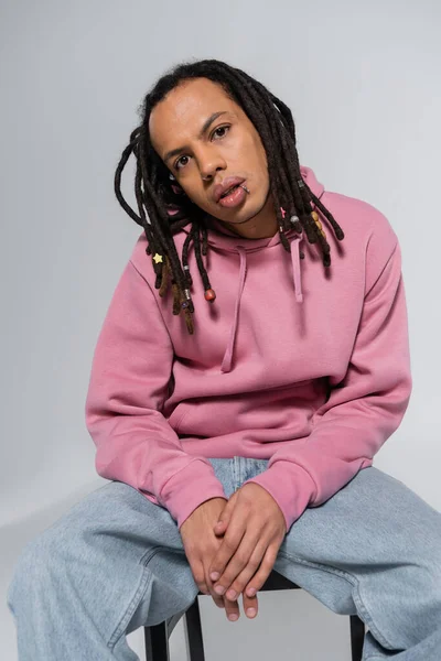 Multiracial and pierced man with dreadlocks looking at camera while sitting isolated on grey — Stock Photo