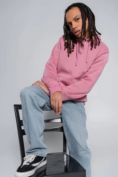 Multiracial and pierced man with dreadlocks looking at camera while sitting on chair back isolated on grey — Stockfoto