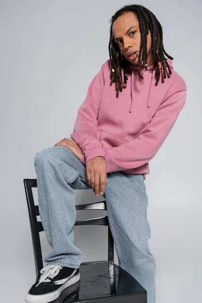 Multiracial and pierced man with dreadlocks looking at camera while sitting on chair back isolated on grey — Stock Photo