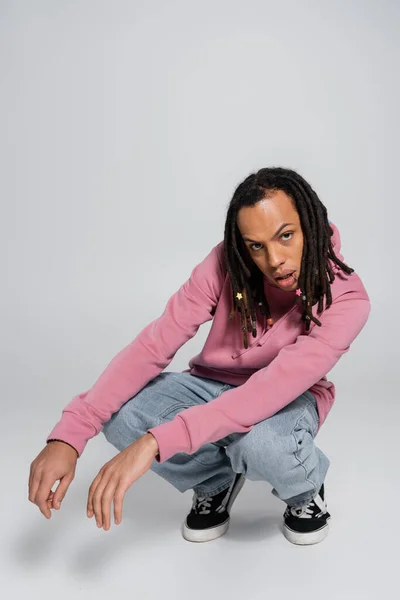 Full length of multiracial man with dreadlocks sitting on haunches and looking at camera on grey — Stock Photo