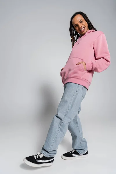 Full length of happy multiracial man in blue jeans and pink hoodie posing with hands in pockets on grey — Stock Photo