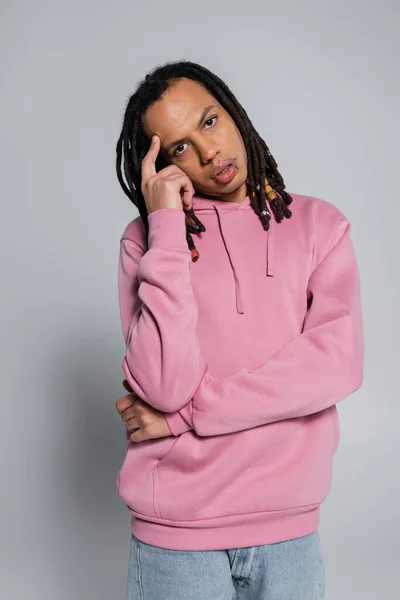 Nachdenklicher multirassischer Mann in rosa Kapuzenpulli blickt in die Kamera auf grau — Stockfoto