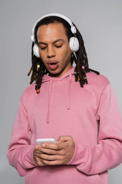 Scioccato uomo multirazziale con dreadlocks utilizzando smartphone durante l'ascolto di musica in cuffie wireless isolato su grigio — Foto stock