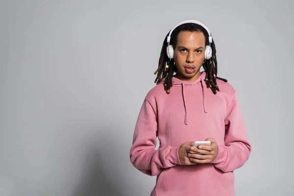 Multiracial man with dreadlocks using smartphone while listening music in wireless headphones on grey background — Stock Photo