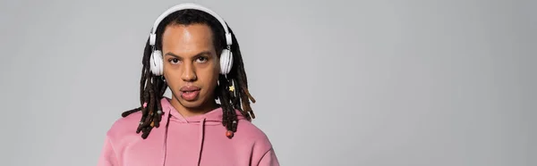 Multiracial man with dreadlocks listening music in wireless headphones isolated on grey, banner — Photo de stock