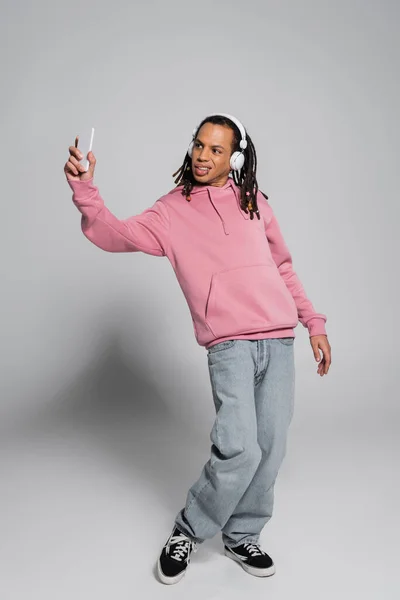 Full length of multiracial man with dreadlocks taking selfie while listening music in wireless headphones on grey — Stock Photo
