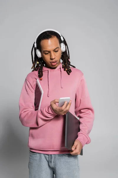 Multiracial man in wireless headphones using smartphone and holding devices on grey — Stock Photo