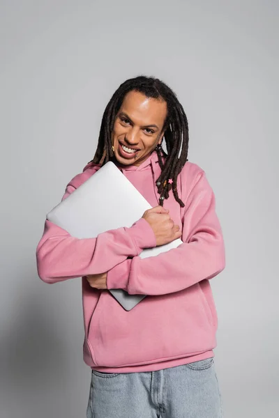 Cheerful and pierced multiracial man in pink hoodie holding laptop on grey — Photo de stock