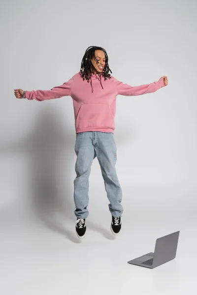 Full length of happy multiracial man in pink hoodie and jeans jumping near laptop on grey — Photo de stock
