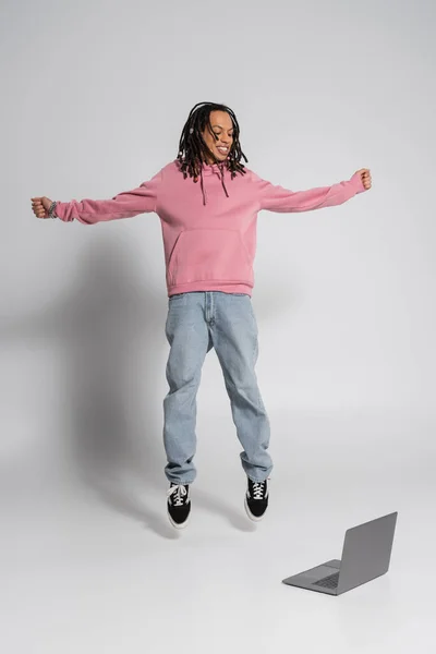 Full length of happy multiracial man in pink hoodie and jeans jumping near laptop on grey — Stock Photo