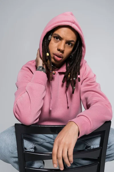 Portrait of tattooed multiracial man in pink hoodie looking at camera while leaning on chair back isolated on grey — Stock Photo