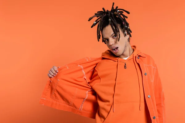 Multiracial man with dreadlocks and opened mouth adjusting denim jacket on coral background — Stock Photo