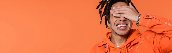 Happy multiracial man with dreadlocks and tattoo covering eyes isolated on coral background, banner — Fotografia de Stock