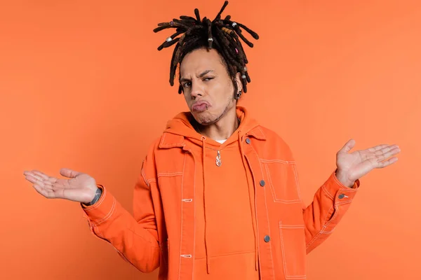 Tattooed and multiracial man with dreadlocks showing shrug gesture isolated on coral background — Stock Photo
