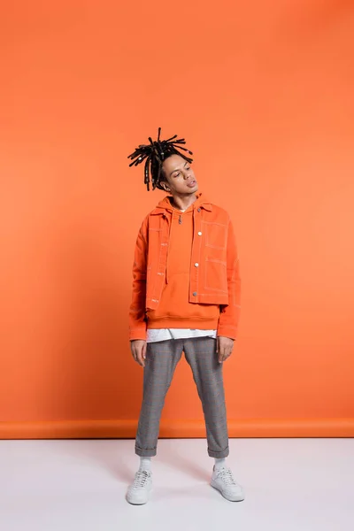 Full length of multiracial man with dreadlocks standing in blue jeans and orange jacket on coral background - foto de stock