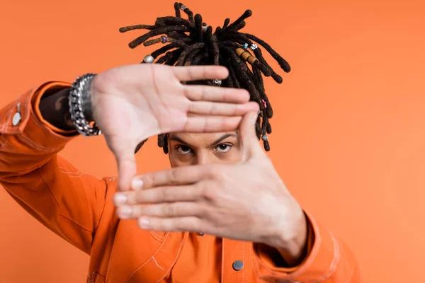 Multiracial man with dreadlocks showing frame gesture while looking at camera isolated on coral background — Stock Photo