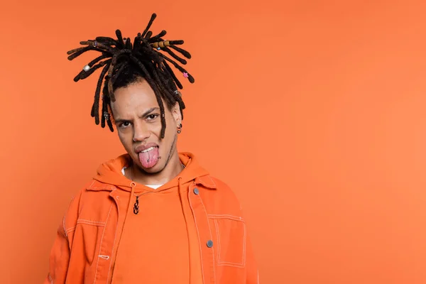 Pierced and multiracial man with dreadlocks looking at camera while sticking out tongue isolated on coral background — Photo de stock