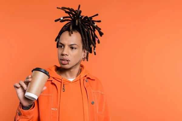 Pensive and multiracial man with dreadlocks holding paper cup with coffee to go isolated on coral background — Stock Photo