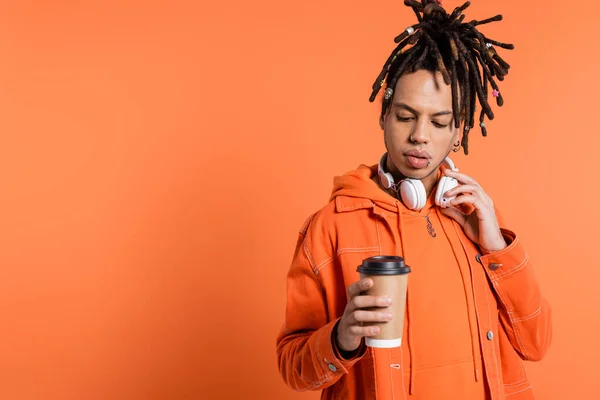 Pierced and multiracial man with wireless headphones looking at paper cup with coffee to go on coral background — Stock Photo