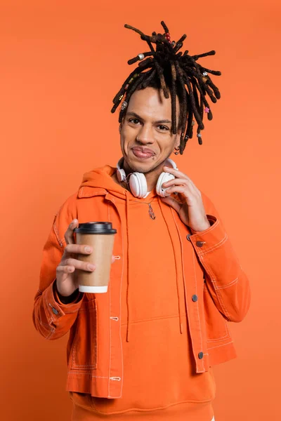 Cheerful and multiracial man with wireless headphones holding paper cup with coffee to go isolated on coral — Stock Photo