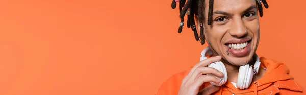 Cheerful and multiracial man with wireless headphones looking at camera isolated on coral background, banner — Fotografia de Stock