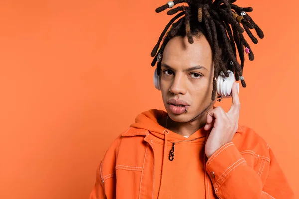 Pierced and multiracial man listening music in wireless headphones while looking at camera on coral background — Stock Photo