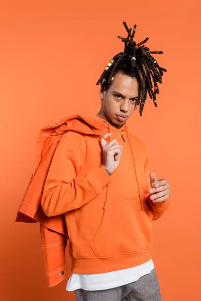 Tattooed and multiracial man in hoodie holding denim jacket isolated on coral background — Stock Photo
