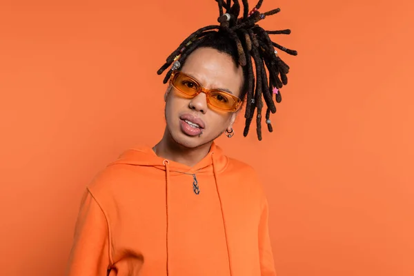 Doubtful multiracial man with dreadlocks and stylish sunglasses looking at camera isolated on coral background — Stock Photo