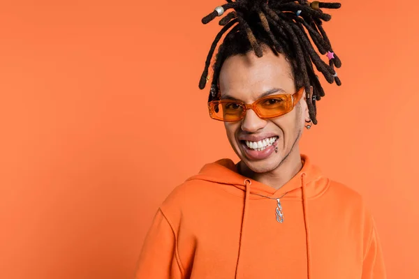 Happy multiracial man with dreadlocks wearing stylish sunglasses isolated on coral background — Stockfoto