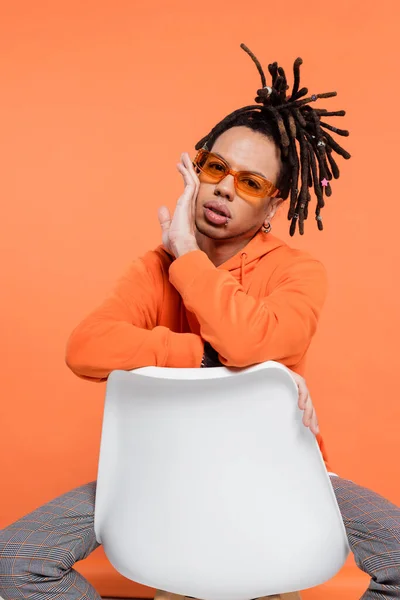 Multiracial man with dreadlocks and sunglasses sitting on chair isolated on coral — Foto stock