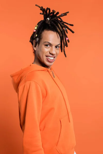 Alegre hombre multirracial con rastas mirando a la cámara y sonriendo aislado en coral - foto de stock