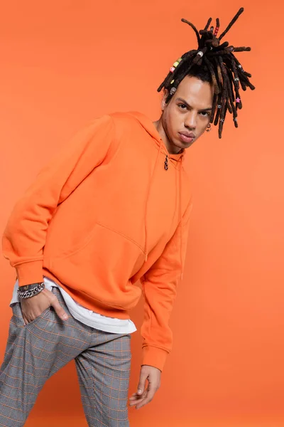 Pierced multiracial man with dreadlocks standing with hand in pocket isolated on coral background — Stock Photo