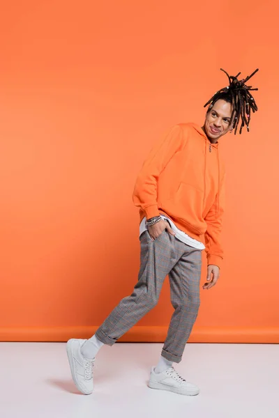 Pleine longueur de sourire homme multiracial avec dreadlocks debout avec la main dans la poche sur fond de corail — Photo de stock