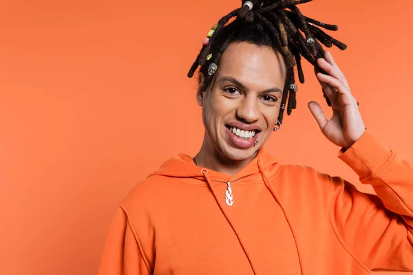 Pleased multiracial man adjusting dreadlocks and smiling isolated on coral — Photo de stock