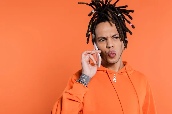 Tattooed multiracial man with dreadlocks pouting lips and talking on cellphone isolated on coral — Stock Photo