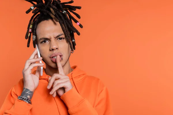 Multiracial man with dreadlocks holding finger near lips and talking on cellphone isolated on coral — Stock Photo