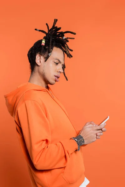 Vista lateral del hombre multirracial tatuado con dreadlocks mensajes de texto en el teléfono inteligente aislado en coral - foto de stock