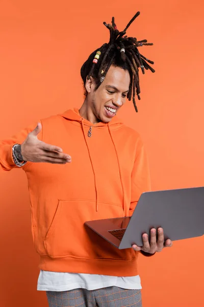 Happy multiracial man with dreadlocks and tattoo pointing at laptop on coral background — Foto stock