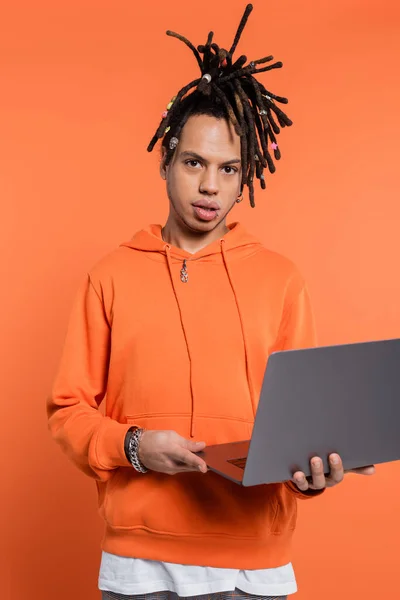 Tattooed multiracial freelancer with dreadlocks holding laptop on coral background - foto de stock